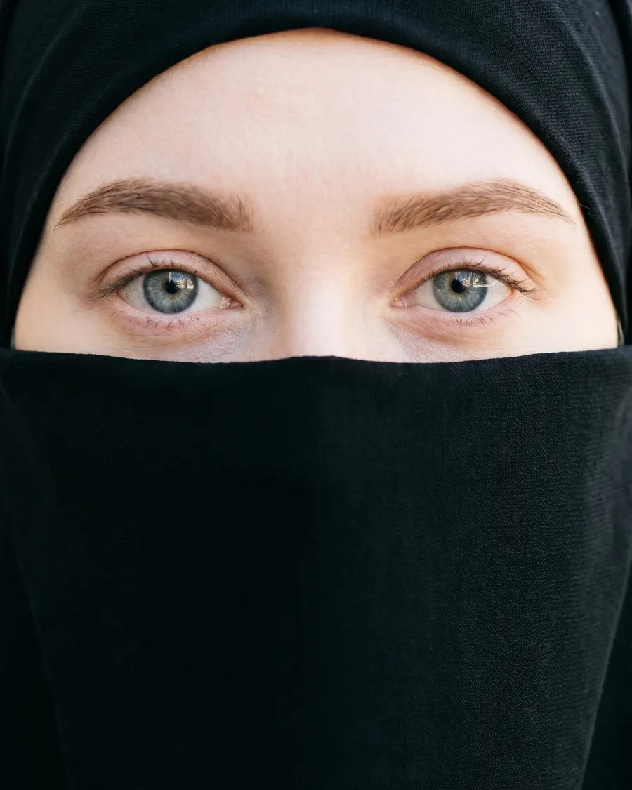 Aisha Moon in a black hijab and niqab with only her eyes visible, photographed against a plain backdrop