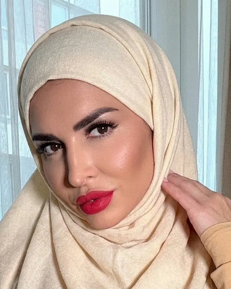 Carolina in a cream hijab with her face visible and red lipstick, posing by a window with sheer curtains in soft daylight