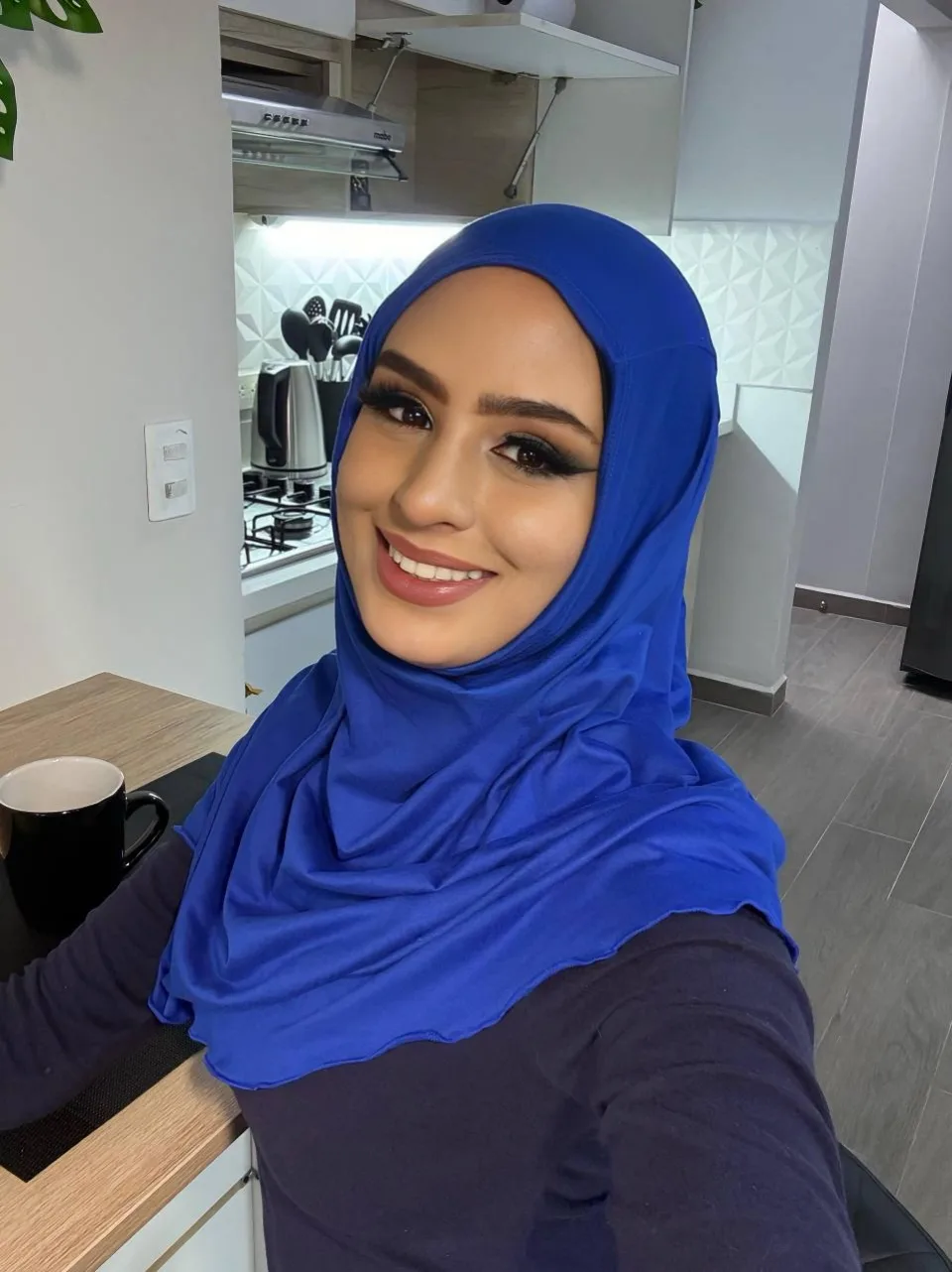 Dalia Fayed in a royal blue hijab and a navy long-sleeved top, smiling in her own kitchen beside a black coffee mug