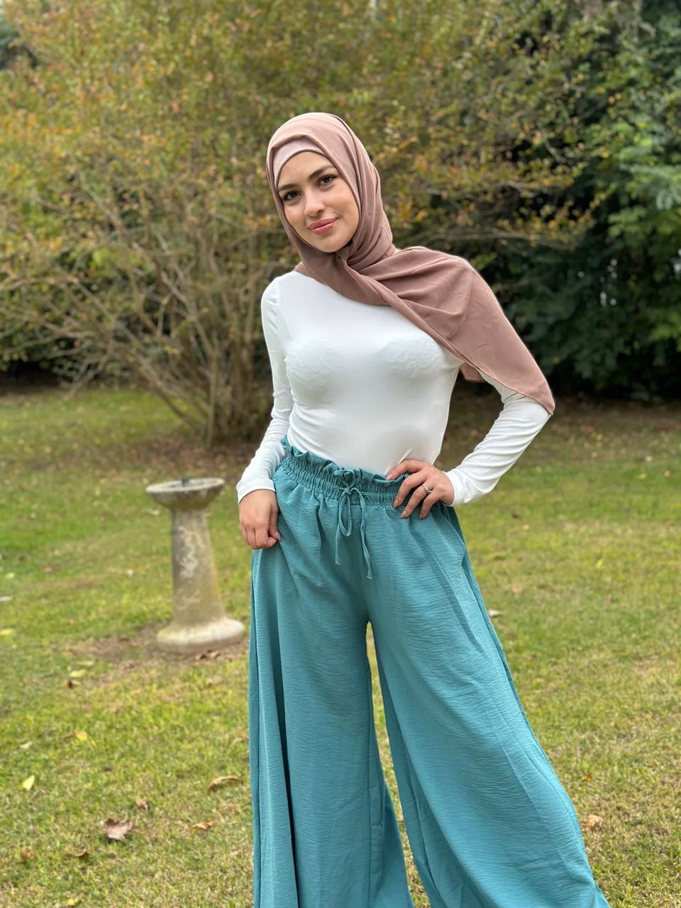 Hessa Khalifa in a taupe hijab, a white long-sleeved top and wide teal trousers, standing on a garden lawn