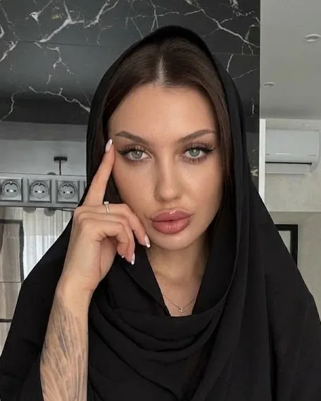 Kate Zadidi in a loosely draped black chiffon hijab with the front of her dark hair showing, one fingertip at her temple, against a black-and-white marble wall