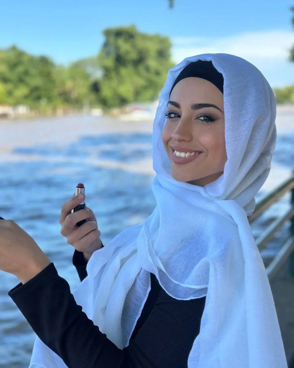 Malika in a white hijab and a black long-sleeved top, photographed beside a river in bright daylight