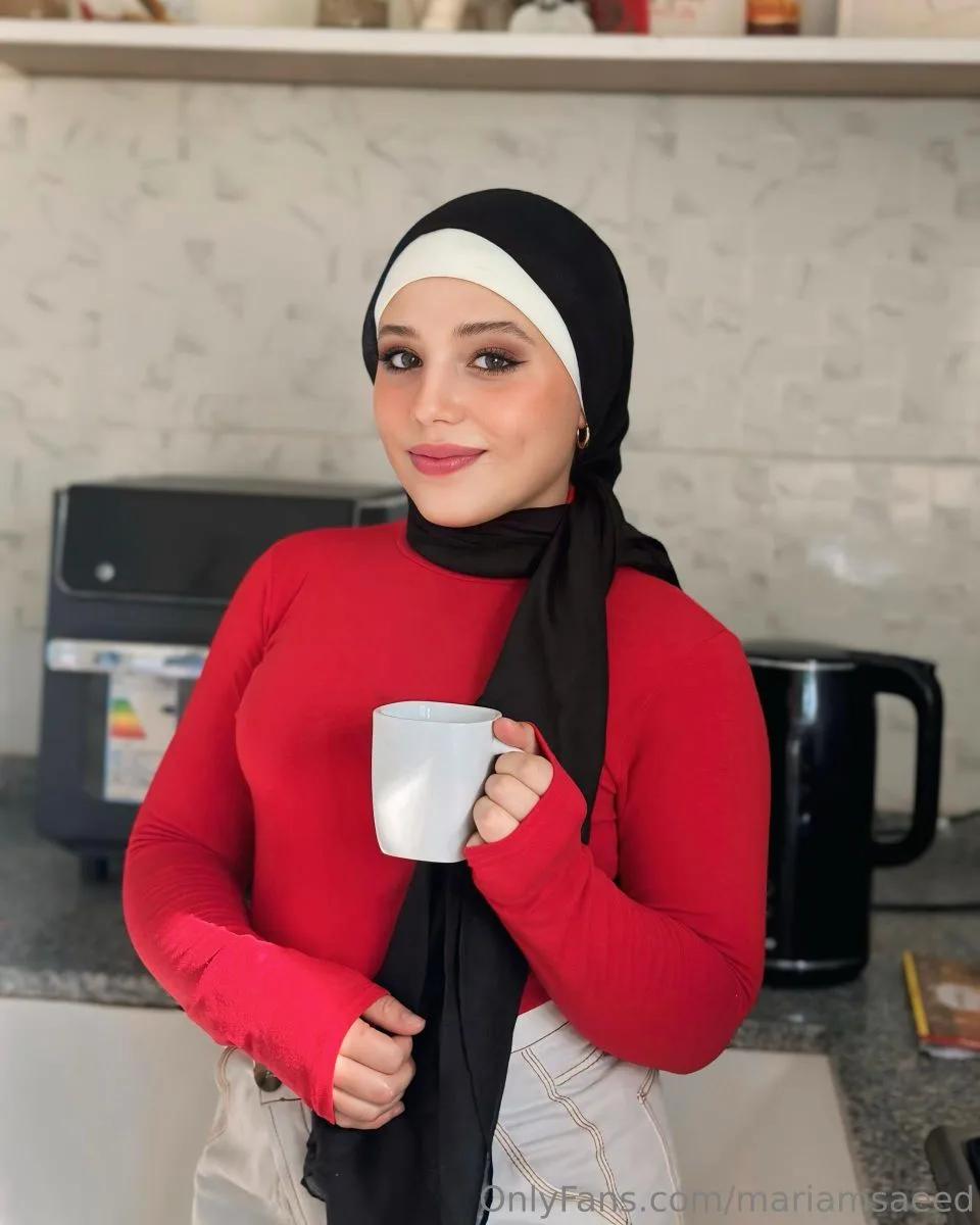 Mariam Saeed in a black and white hijab and a red long-sleeved top, holding a mug in a plain kitchen