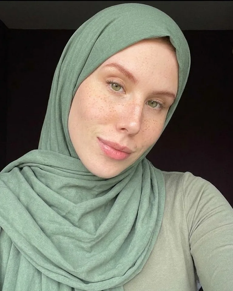 Nasty Darydova in a sage green jersey hijab and a matching long-sleeved top, freckled face and green eyes visible against a dark wall